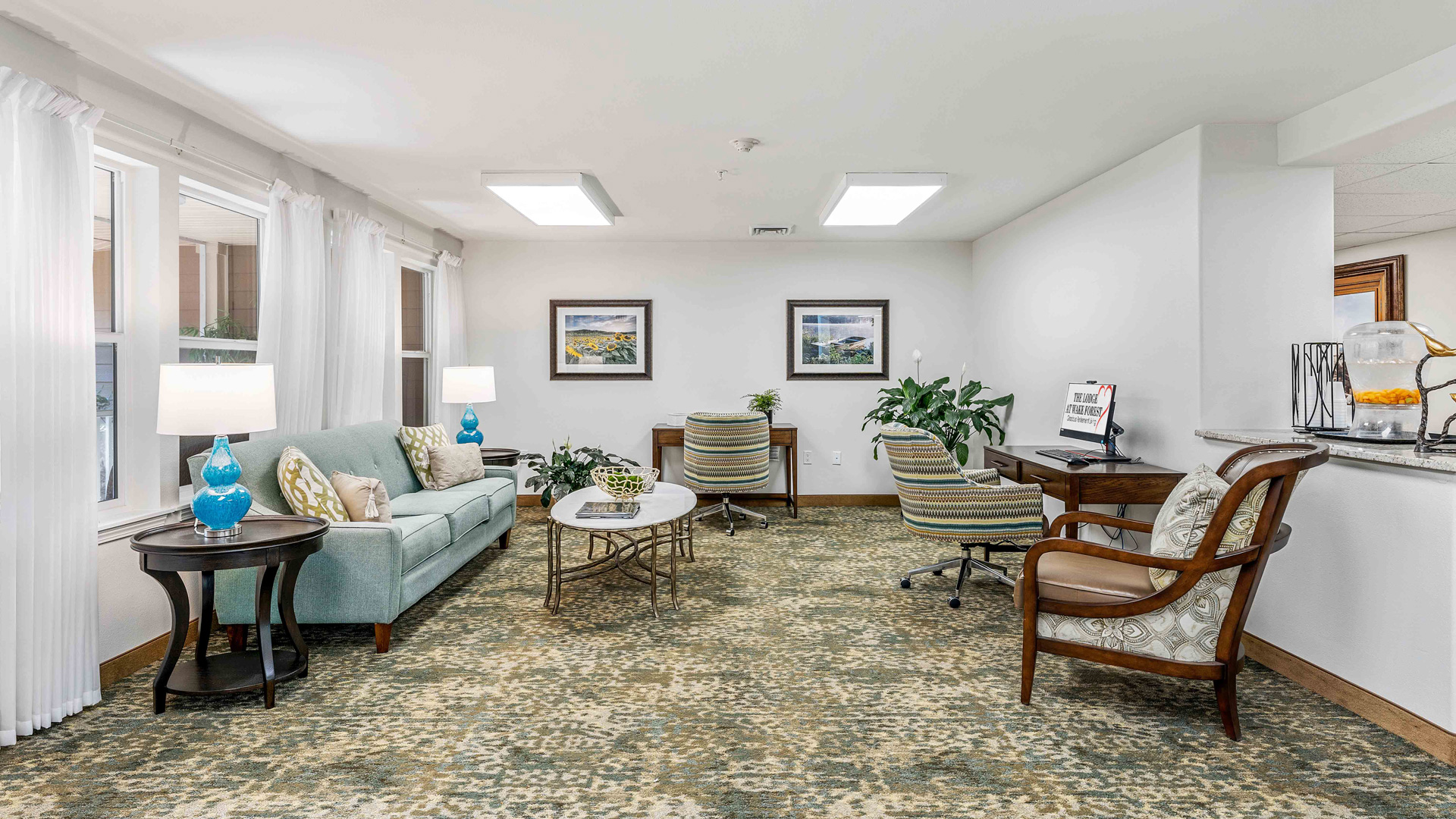 A cozy, well-lit living room with a sofa, armchairs, side tables with lamps, desk, plants, and framed artwork on the wall. Large windows let in natural light, and a refreshment area is on the right.