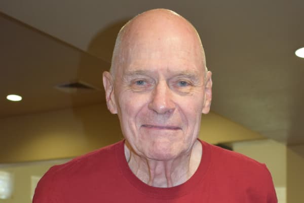 A bald elderly man is smiling gently at the camera. He is wearing a red t-shirt, and the background is a softly lit indoor setting with neutral-colored walls.