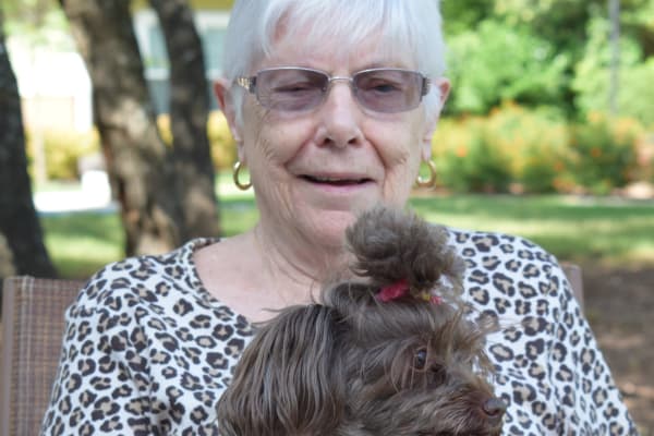 An elderly woman with short white hair and glasses sits outdoors, wearing a leopard print top and smiling. She holds a small, fluffy dog with a brown coat and a tuft of fur tied up on its head. The background includes trees and greenery.