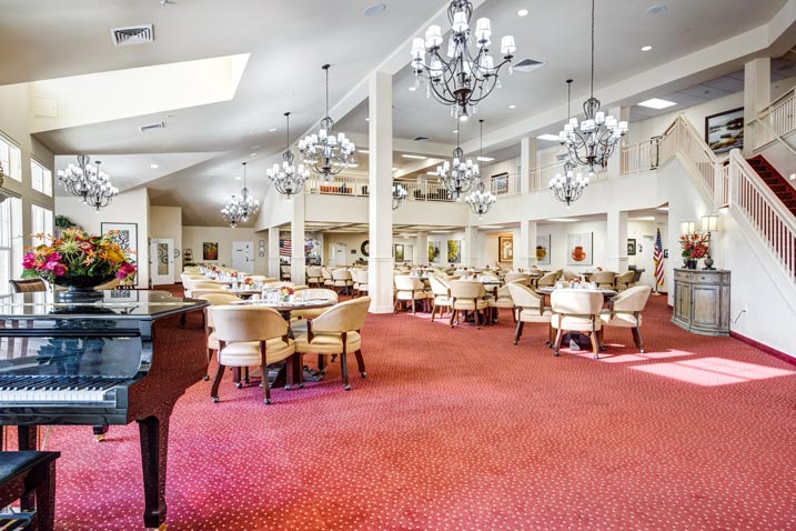 A spacious dining area with red carpet, cream-colored chairs, and round tables. Chandeliers hang from the high ceiling, and there's a grand piano with a flower vase in the foreground. Stairs lead to an upper level, and large windows let in natural light.