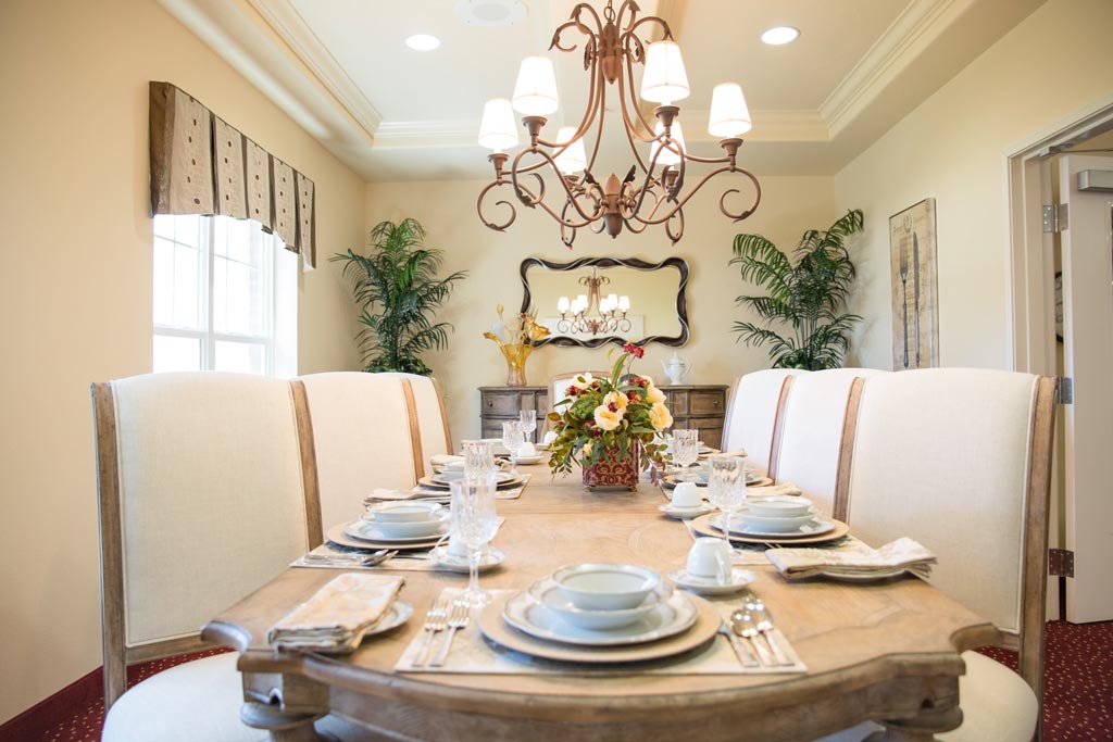 A dining room with a wooden table set for a meal, featuring plates, glasses, and cutlery arranged neatly. High-back upholstered chairs surround the table. A chandelier hangs above, and a mirror, two potted plants, and art decor are visible in the background.