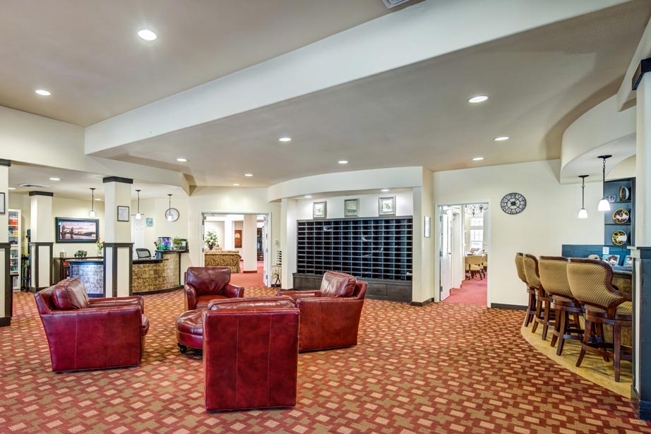 Cozy lobby area with red leather chairs and a round coffee table on a patterned carpet. The room includes a reception desk, mail cubbies, and a bar area with high-back chairs. Decor includes framed pictures and wall art. Warm, inviting atmosphere.