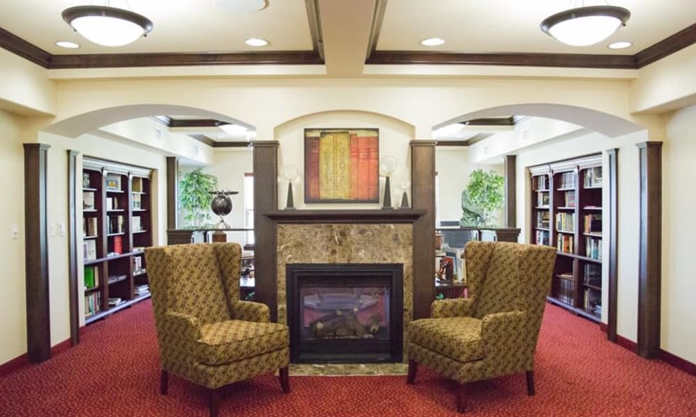 A cozy library room features two patterned armchairs facing a fireplace with a wooden mantle. A bookshelf filled with books is built into each side wall, and large windows allow natural light. Above the fireplace is a framed artwork. The room has a red carpet.