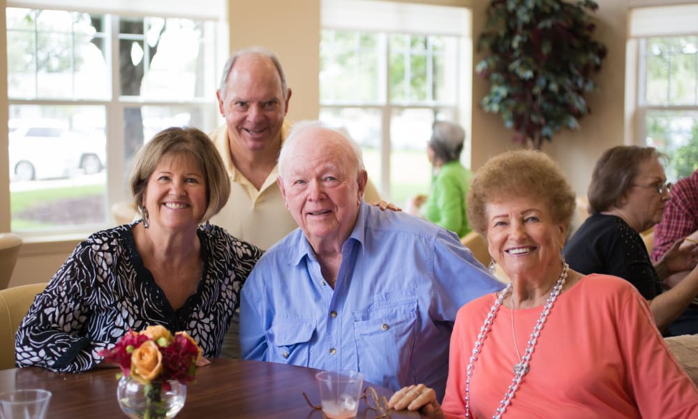 Four elderly people are seated around a table, smiling at the camera. Two are men, one in a light shirt and one in a blue shirt, and two are women, one in a black and white top and the other in an orange top and necklace. A vase of flowers sits on the table.