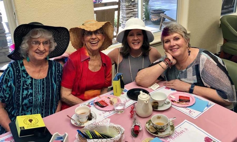 Four women smile while sitting at a table for an afternoon tea party. They are wearing hats and have various tea accessories on the table, including tea cups, a teapot, and plates with pastries. The background includes large windows and greenery outside.