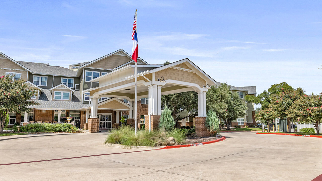 A large, multi-story building with a covered entrance, brick accents, and landscaped greenery. Two flagpoles display the American and Texas flags near the driveway, with clear skies above.