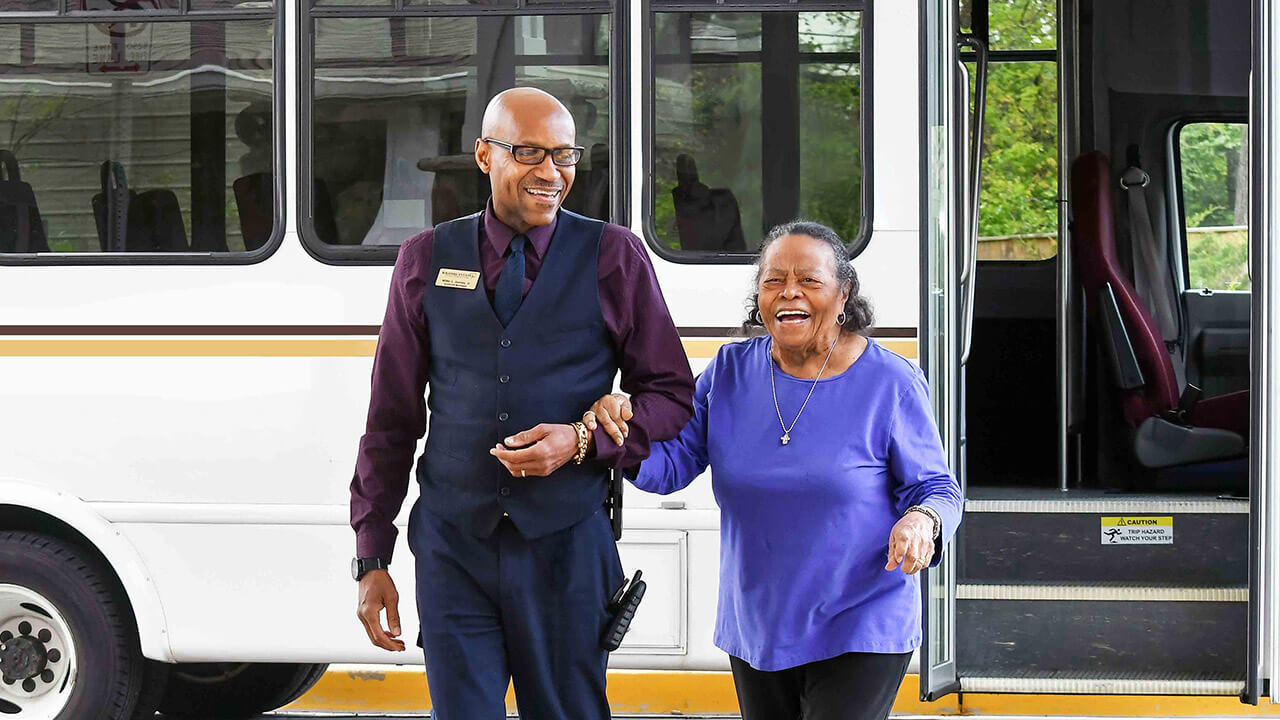 A smiling man in a vest and name tag walks arm-in-arm with a laughing older woman in a purple top, guiding her off a white shuttle bus with open doors.