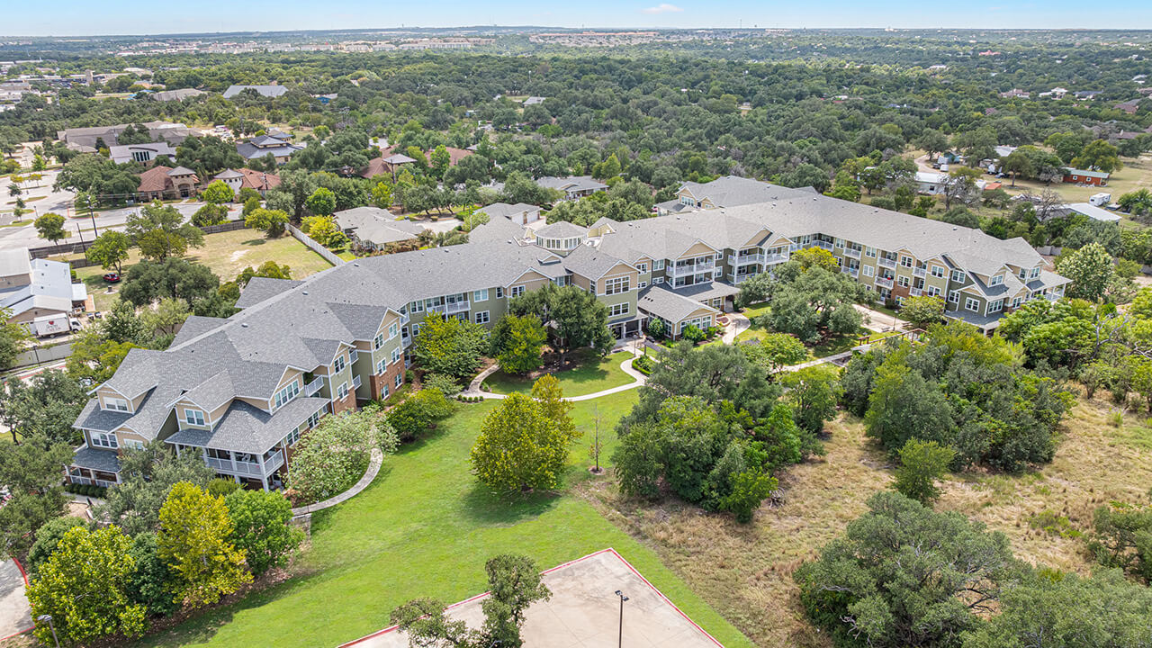 Aerial view of a large, multi-story residential building surrounded by green trees and grassy areas, with a suburban landscape and scattered houses in the background.