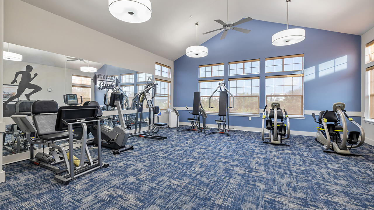 A bright gym room with blue carpet, large windows, exercise machines, and fitness equipment. A wall mural of a running person is reflected in a mirror on the left side. Ceiling fans and lights hang from the white ceiling.