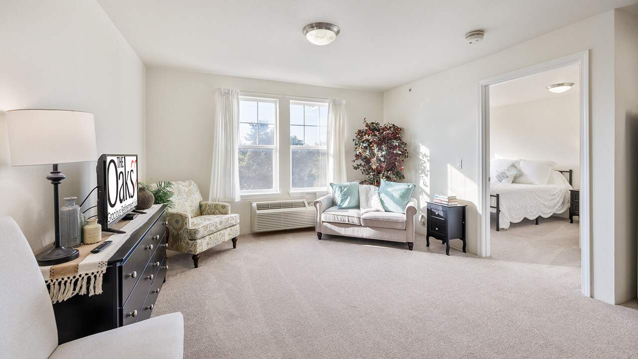 A bright, carpeted living room with a TV, armchair, sofa, dresser, potted plant, and large window. An open doorway leads to a bedroom with a neatly made bed and white bedding.