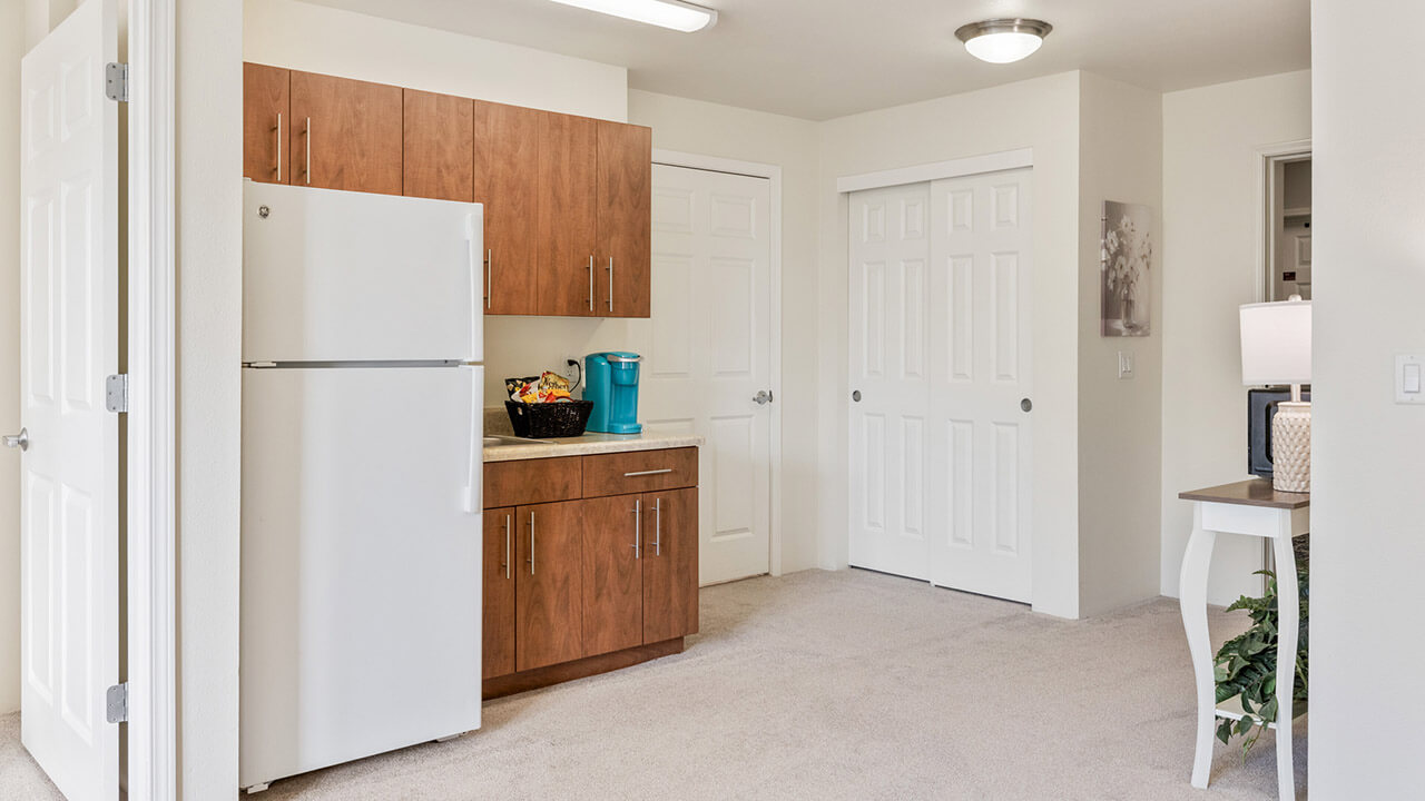 A small kitchen with brown cabinets, a white refrigerator, beige countertop, and a teal coffee maker. The space has carpeted flooring, white walls, and several closed white doors. A small table with a plant is on the right.