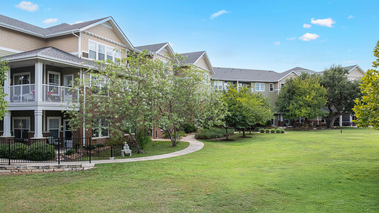 Two-story apartment buildings with balconies overlook a large, well-maintained grassy lawn with trees and a winding sidewalk on a sunny day.