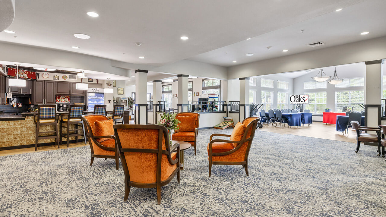 A spacious, well-lit lounge area with four orange armchairs around a small table, a bar with stools to the left, and tables with chairs by large windows in the background. The décor is modern and inviting.