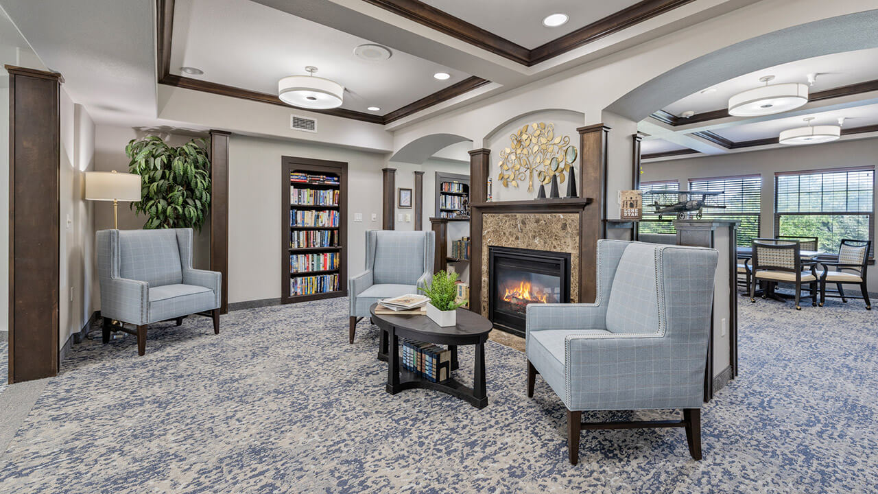 Modern lounge area with blue armchairs, a small coffee table, fireplace, bookshelf, and decorative wall art. Large windows bring in natural light, and a dining area is visible in the background.