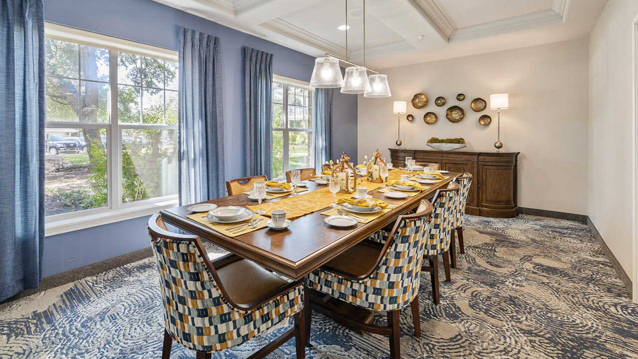 A dining room with a long wooden table set for eight, patterned chairs, blue curtains, large windows, a sideboard with lamps, and decorative wall plates. The room has bright lighting and a patterned carpet.