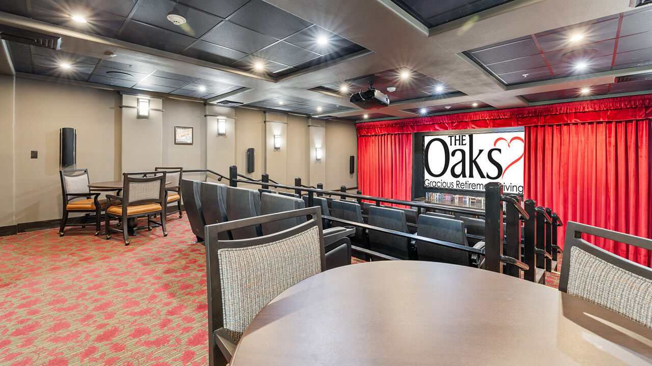 A small theater room with tiered seating, tables and chairs, red carpet, and a large screen displaying "The Oaks Gracious Retirement Living" in front of closed red curtains.