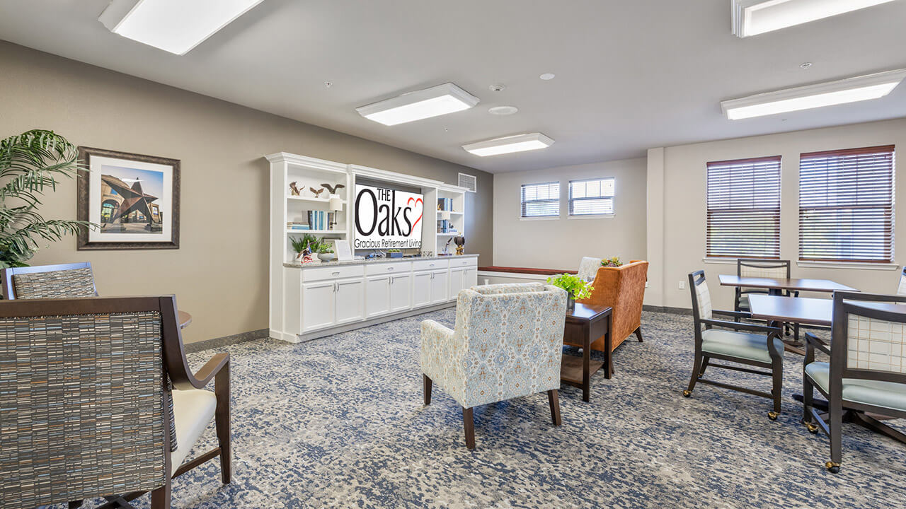 A bright, modern lounge with patterned blue carpet, various armchairs, a white shelving unit with "The Oaks" sign, framed art, potted plants, and large windows with wooden blinds letting in natural light.