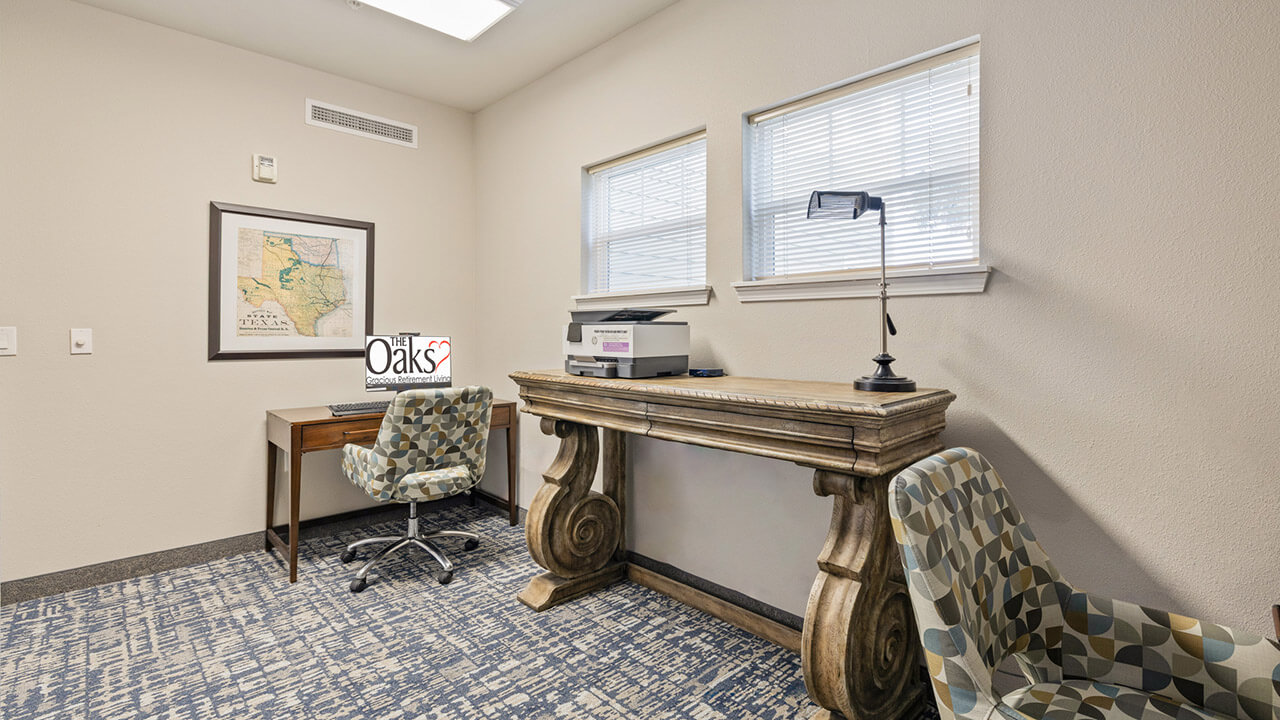 A small office with a patterned blue carpet, a wooden desk with a printer and lamp, a cushioned chair, a second desk with a computer, and a map on the wall, lit by two windows.