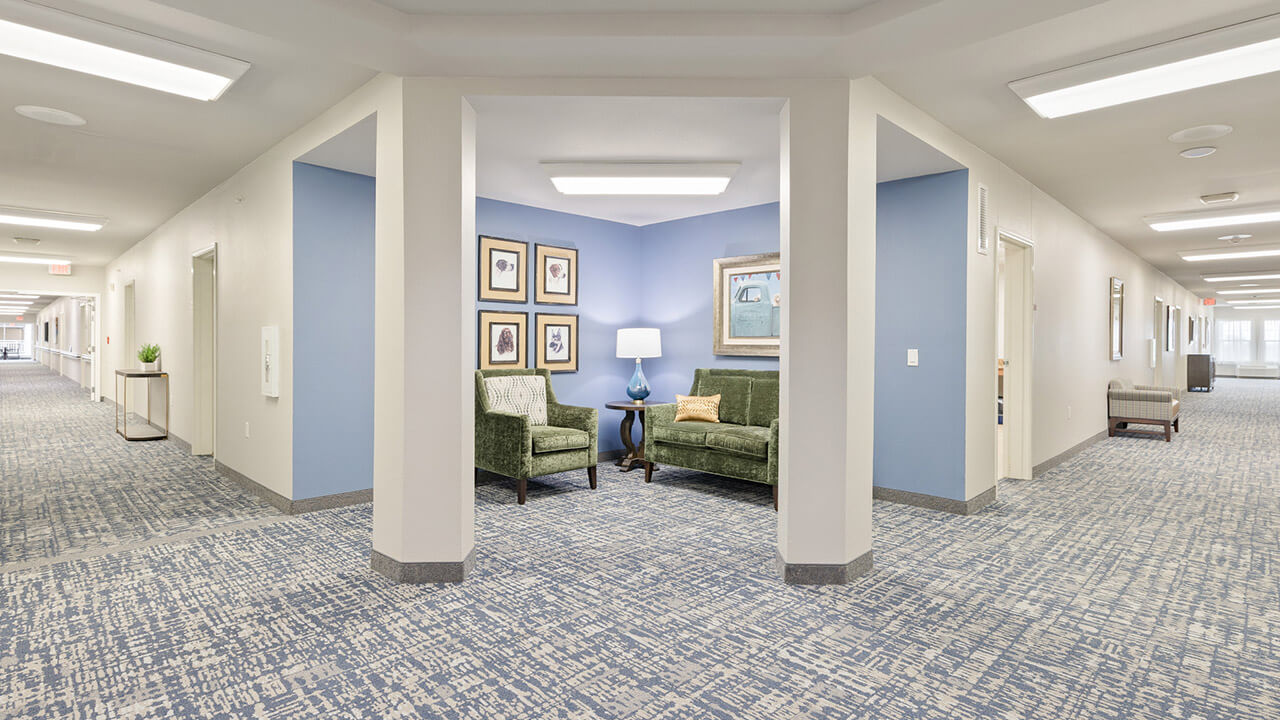 A clean, modern hallway with patterned blue carpet, white walls, and blue accent walls. A cozy seating nook with two green chairs, a lamp, and framed art is set in the middle of the hall, with doors to rooms on either side.