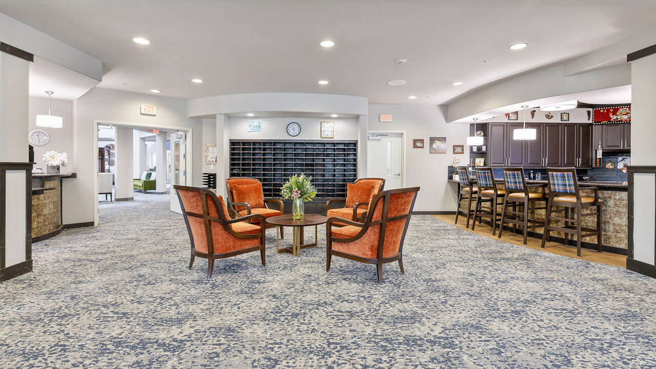 A spacious, well-lit lounge with four orange chairs around a coffee table in the center, a blue patterned carpet, a wall of mailboxes, and a bar area with high chairs in the background.