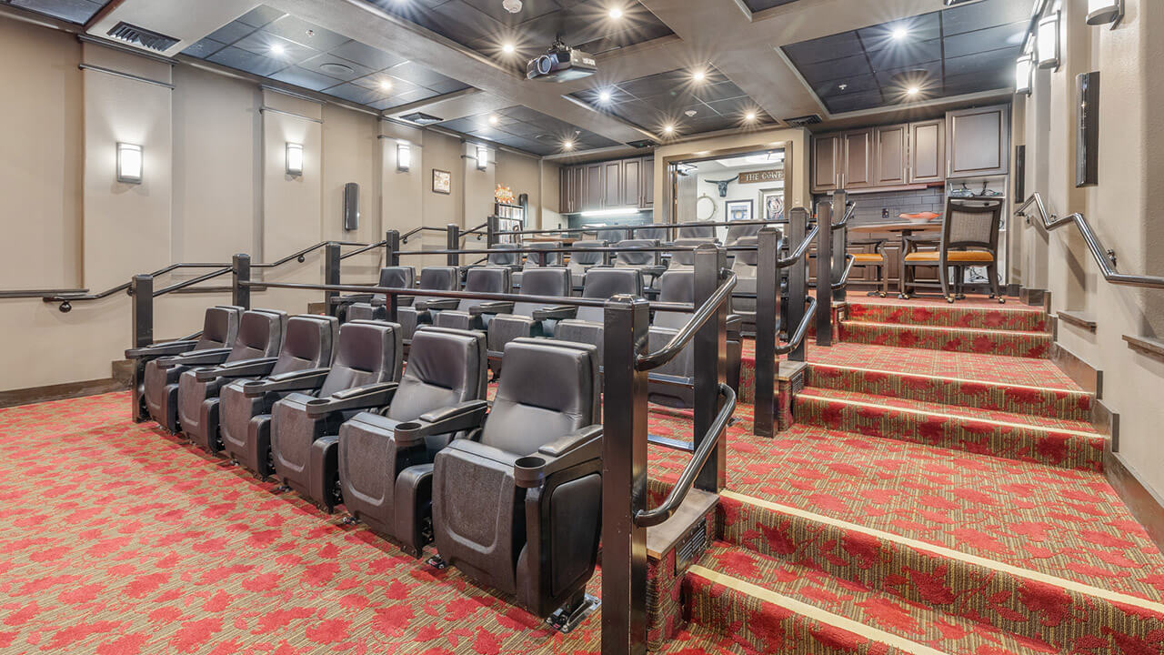 A small private theater with tiered rows of black cushioned seats, red patterned carpet, beige walls, and wall-mounted lights; a countertop and chairs are visible at the back of the room.