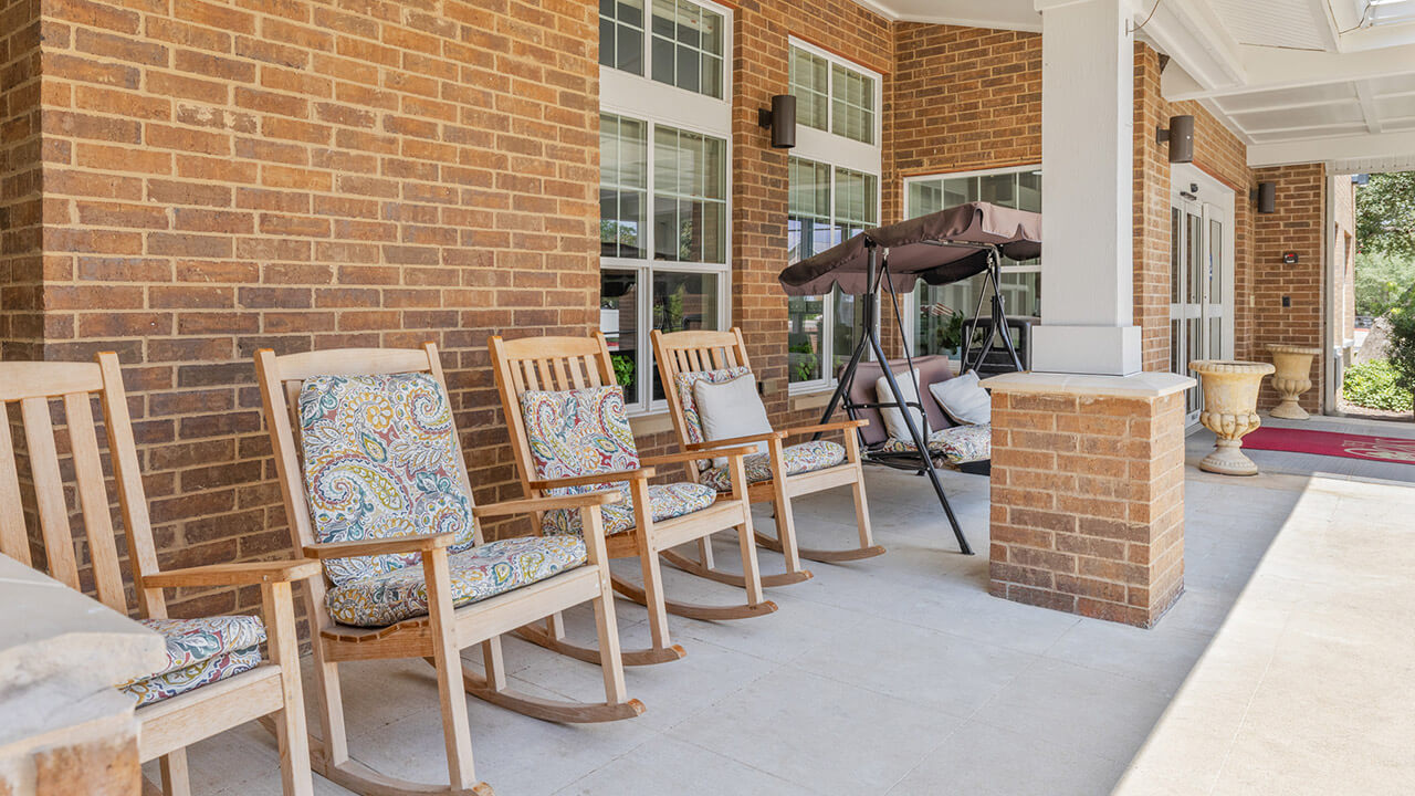 A row of wooden rocking chairs with patterned cushions sits on a covered porch with brick walls and large windows. A cushioned porch swing is visible in the background.