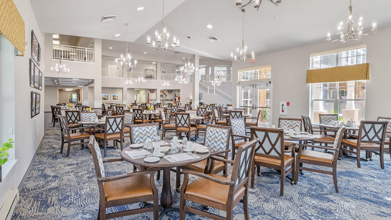 A spacious, well-lit dining room with neatly set round tables and wooden chairs, chandeliers hanging from the ceiling, large windows, and a modern, elegant decor.