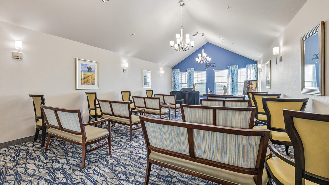 A small, elegant meeting room with rows of cushioned chairs facing a table and two chairs at the front, decorated with wall art, mirrors, blue accent wall, chandelier, and large windows with blue curtains.