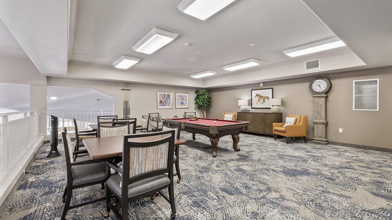 A spacious recreation room with a pool table, several tables and chairs, wall art, a large clock, an armchair, and indoor plants, all under bright ceiling lights and a patterned carpet.