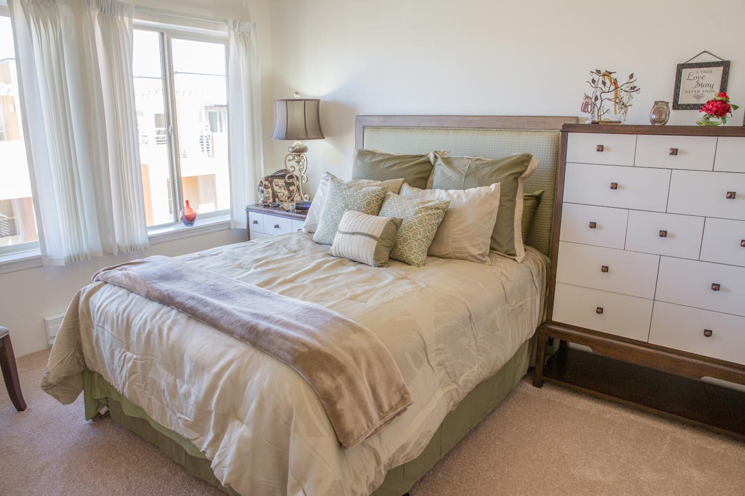 A cozy bedroom featuring a neatly made bed with multiple pillows and a beige comforter. There's a nightstand with a lamp and decor near the window. A dresser with drawers is against the right wall, topped with various decorative items. Light fills the room through the window.