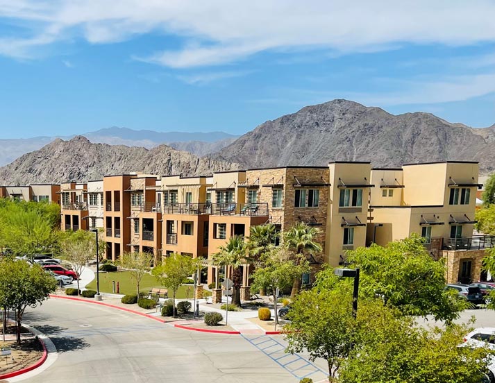 Modern, multi-story apartment complex with desert landscaping and mountainous backdrop. The buildings are cream-colored with stone accents and balconies. A winding road with parked cars runs in front, and the sky is clear with a few scattered clouds.