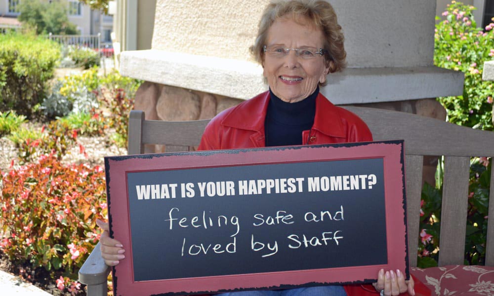 An elderly woman wearing a red jacket and glasses is sitting on an outdoor bench, smiling and holding a sign that reads, 