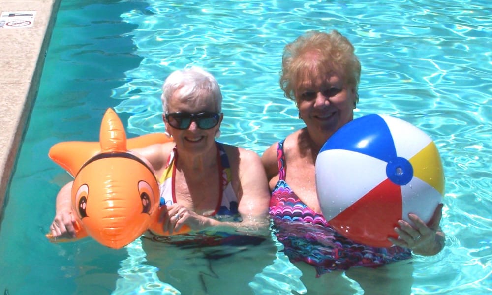 Two elderly women are standing in a swimming pool. Both are smiling and wearing swimsuits. One woman has a bright orange inflatable toy shaped like a fish, and the other is holding a colorful beach ball. The water is clear and the sun is shining.