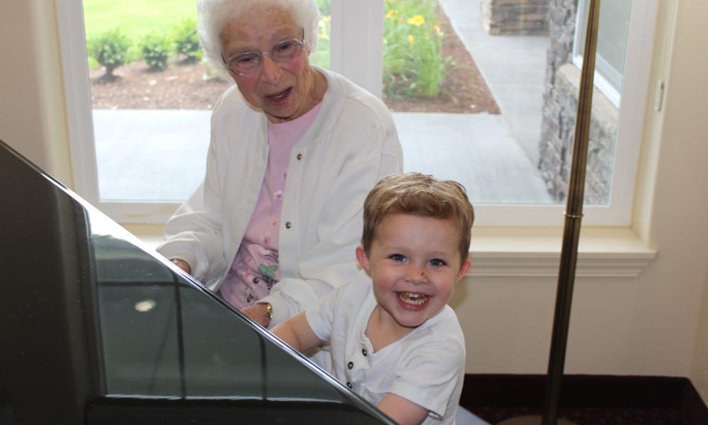 An elderly woman with white hair and glasses sits beside a young boy playing piano keys, both smiling. The setting appears to be indoors near a window overlooking a garden.