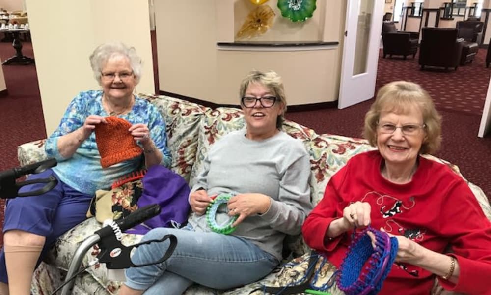 Three elderly women are sitting on a couch in a warmly lit room, smiling and knitting colorful items. They appear relaxed and happy, enjoying each other's company. Two walking aids are visible in front of them, and the room's decor includes colorful wall art.