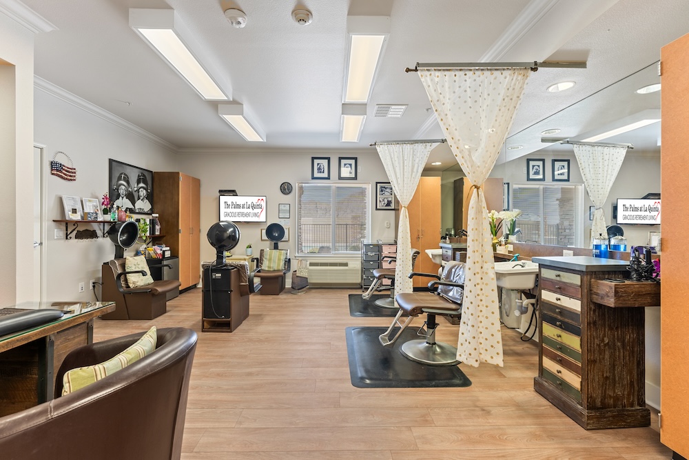 A spacious hair salon with wooden flooring and multiple workstations. Each station has a styling chair, mirrors, and hairdressing equipment. Decorative curtains divide the space, and framed pictures adorn the walls. Bright overhead lighting illuminates the room.