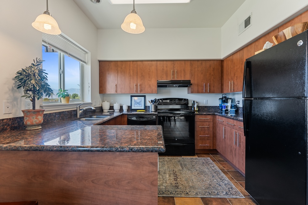 A modern kitchen with wooden cabinets, black appliances, and granite countertops. Two pendant lights hang above the breakfast bar. A window lets in natural light, and a small plant decorates the space. A rug lies on the tile floor.