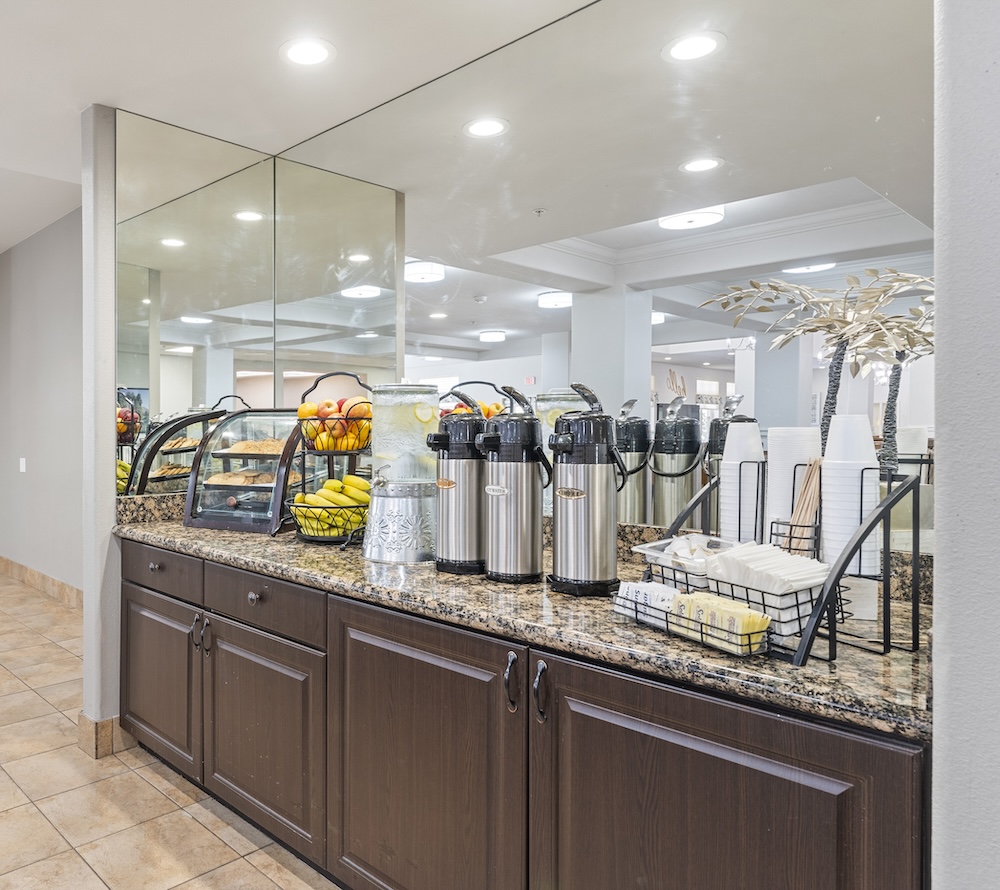 A hotel breakfast bar with coffee thermoses, a fruit basket with bananas and apples, a dispenser filled with lemonade, stacks of cups, and a basket containing condiments. The counter is made of granite, and large mirrors are above it.