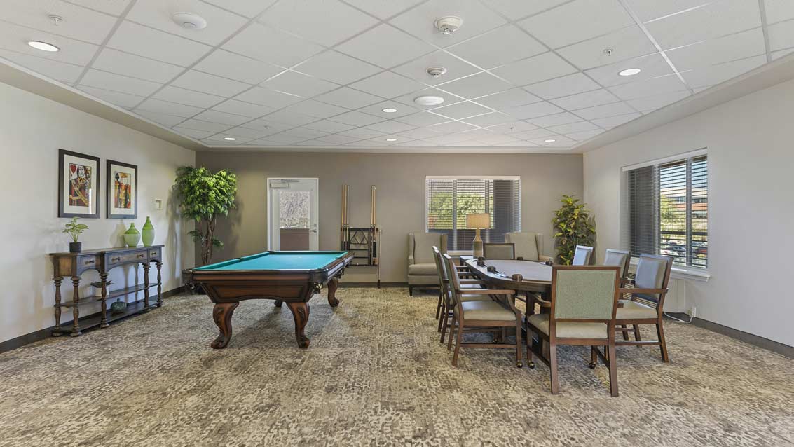 A well-lit recreation room featuring a pool table, a card table with multiple chairs, framed artwork on the walls, and large windows allowing natural light. The room also includes decorative plants and a console table with green vases.