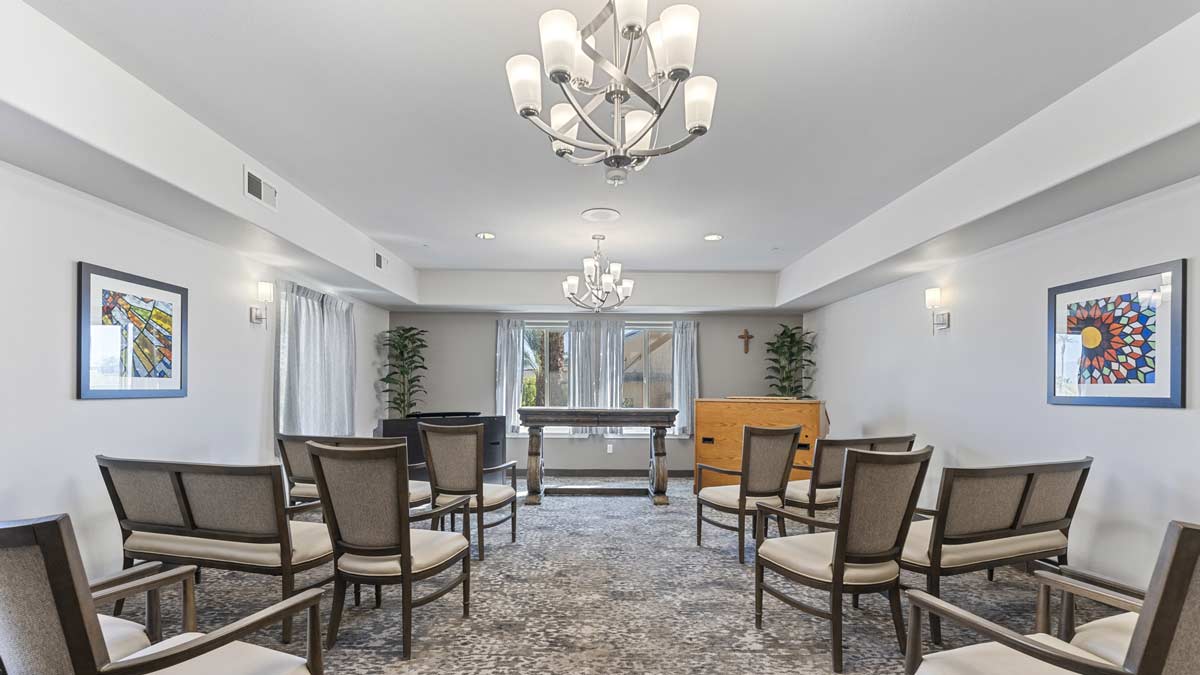 A well-lit, modern funeral home chapel with an arrangement of chairs facing an altar. The room has a chandelier, framed artwork on the walls, a cross in the corner, and large windows letting in natural light. The carpet has a subtle pattern, and there are green plants.