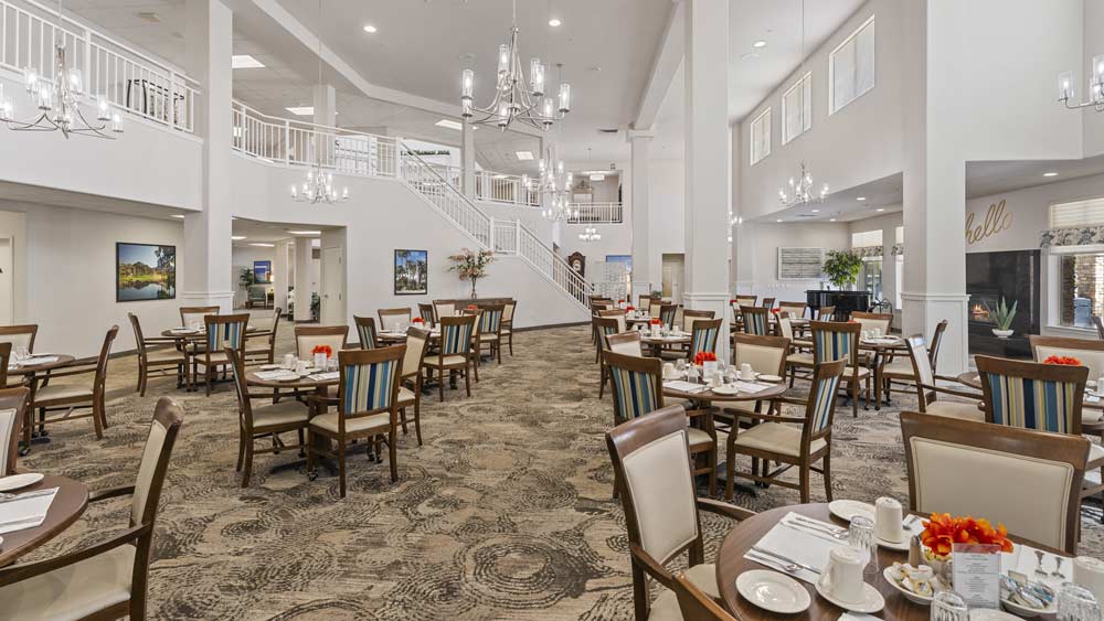 A spacious dining area with multiple tables and chairs, set for a meal with white tablecloths and red flowers as centerpieces. The room features high ceilings, elegant chandeliers, and a double staircase leading to an upper level. Artwork adorns the white walls.