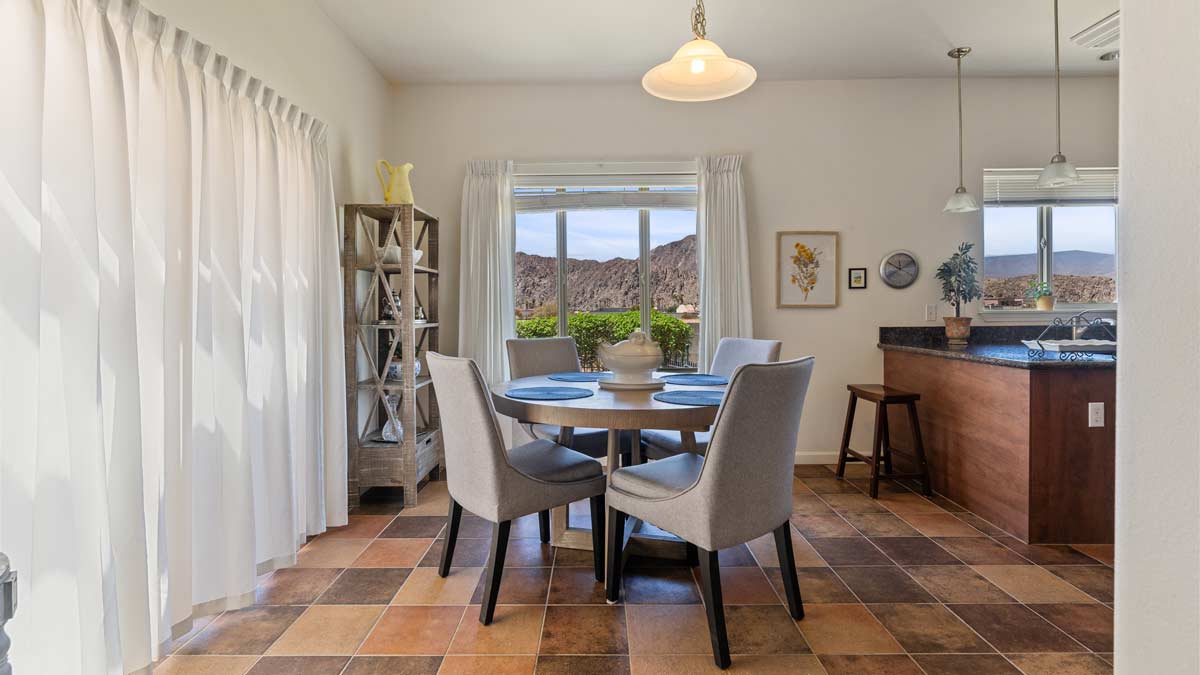 A bright dining area with a round table set for four, featuring gray upholstered chairs on a tiled floor. Sunlight streams through large windows with white curtains, revealing a scenic mountain view. A small modern kitchen is visible to the right.