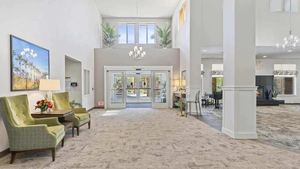 A spacious lobby with high ceilings, modern decor, and plenty of natural light. Two green upholstered chairs with a small wooden table and flowers sit to the left. A reception desk and more seating areas are visible in the background. Large double glass doors lead outside.