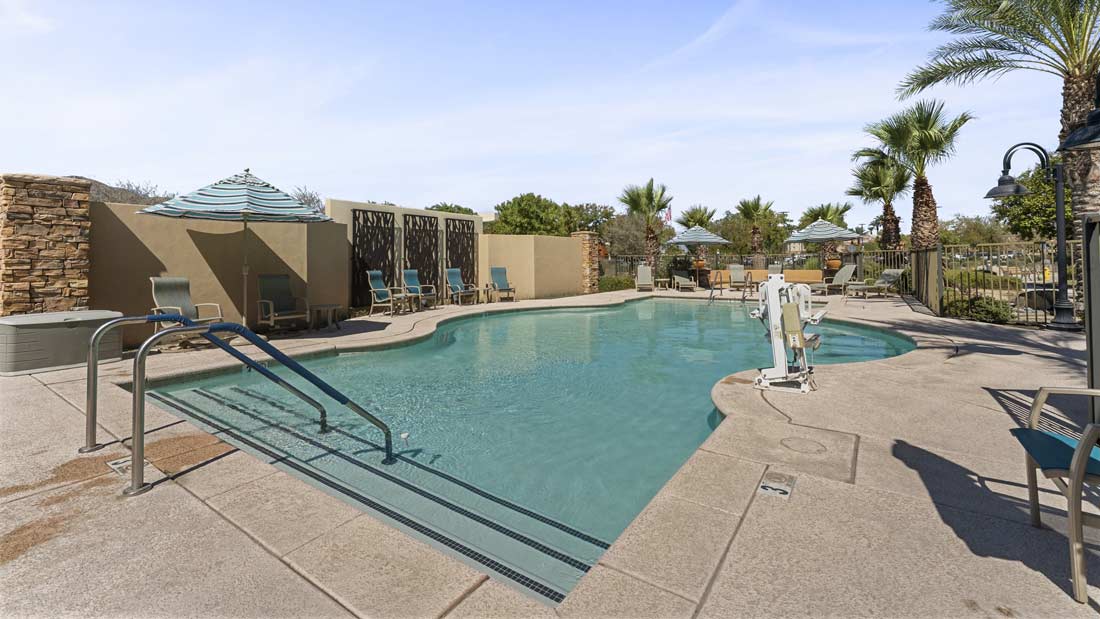 An outdoor swimming pool features steps with railings on the left and a pool lift on the right. Surrounding the pool are sun loungers with umbrellas, a few palm trees, and a stone wall. The sky is clear with some light clouds.