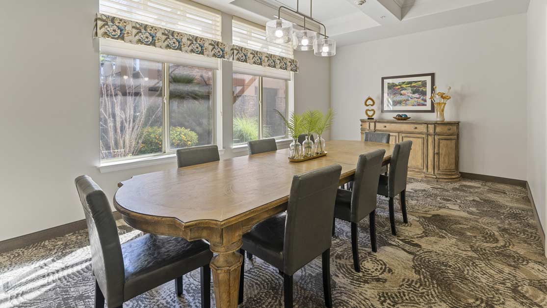 A dining room with a long wooden table surrounded by eight black upholstered chairs. Large windows with patterned valances let in natural light. A wooden sideboard with decorative items and a framed picture is against the back wall. Carpet features a swirling design.