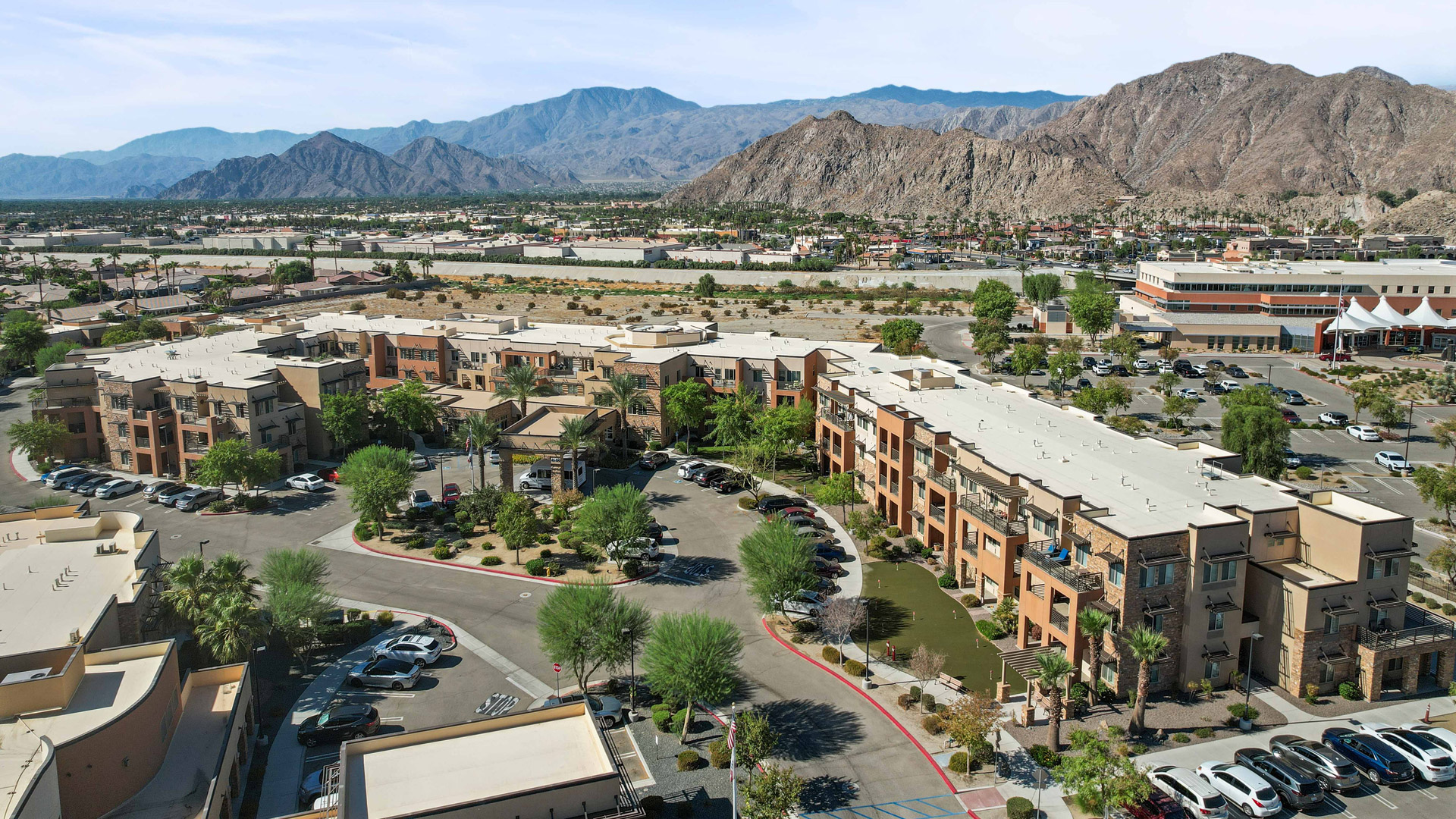Aerial view of a modern apartment complex with tan buildings, green trees, and parked cars, set against a backdrop of mountains and a sprawling cityscape under a clear sky.
