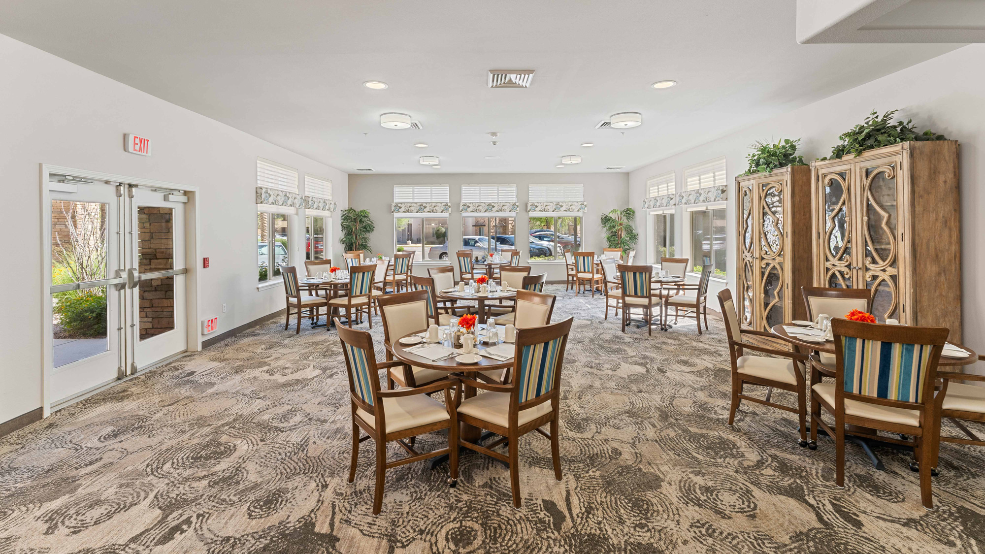 A bright, spacious dining room with round tables set for meals, striped cushioned chairs, large windows letting in natural light, and decorative cabinets along the walls.