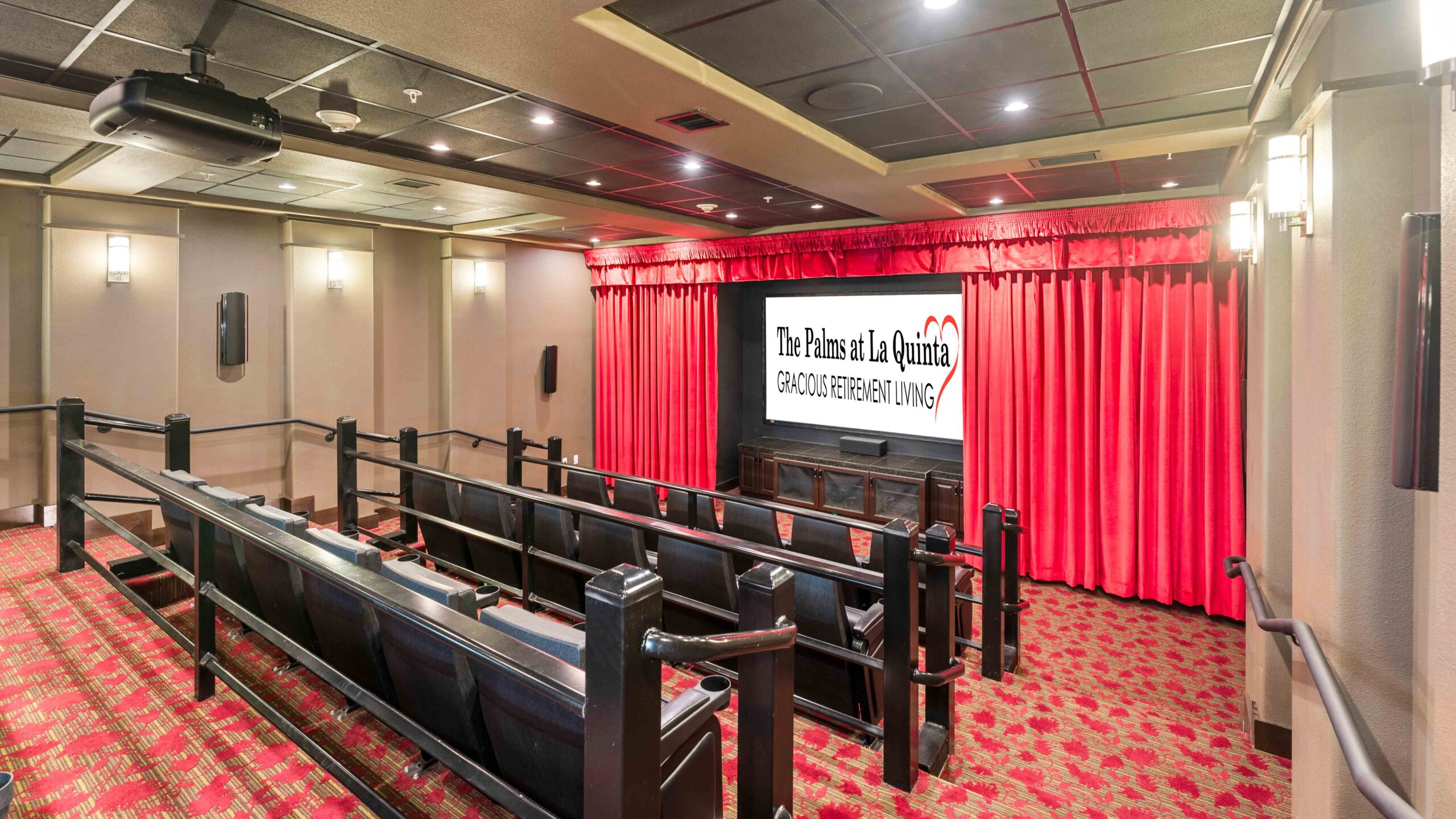 A small theater room with tiered seating, black chairs, red carpet, and red stage curtains. A screen at the front displays "The Palms at La Quinta Gracious Retirement Living.