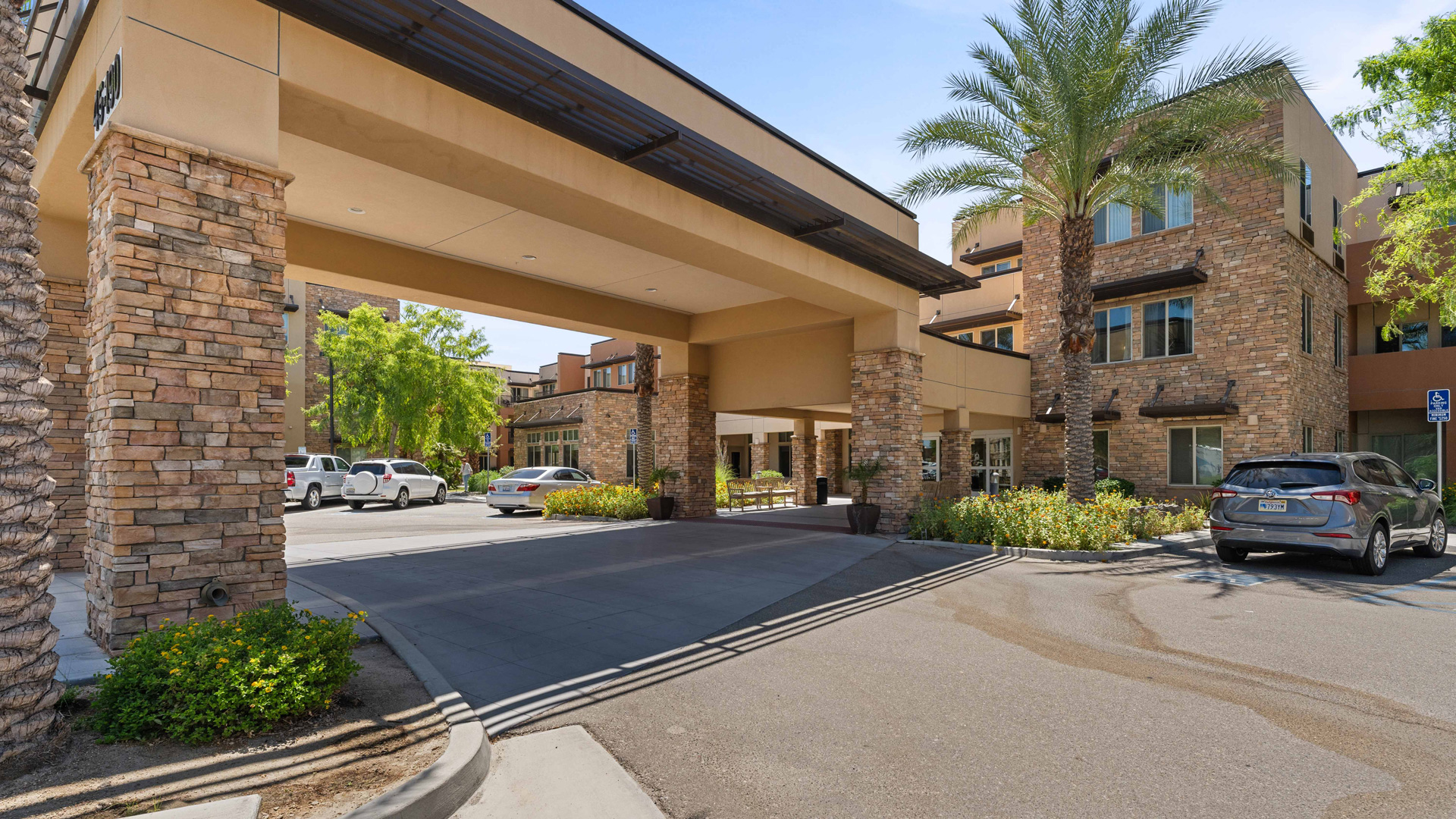 A modern building with stone and beige accents features a covered driveway entrance, surrounded by palm trees, shrubs, and parked cars on a sunny day.