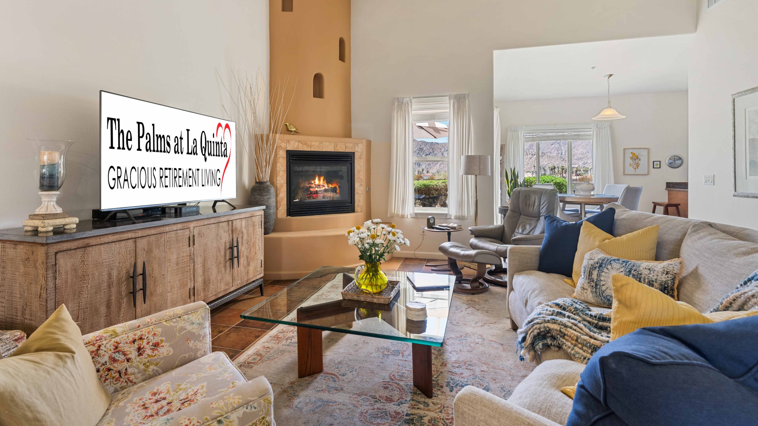 A bright living room with a fireplace, a TV displaying "The Palms at La Quinta Gracious Retirement Living," a glass coffee table with flowers, and comfortable seating with yellow and blue pillows.